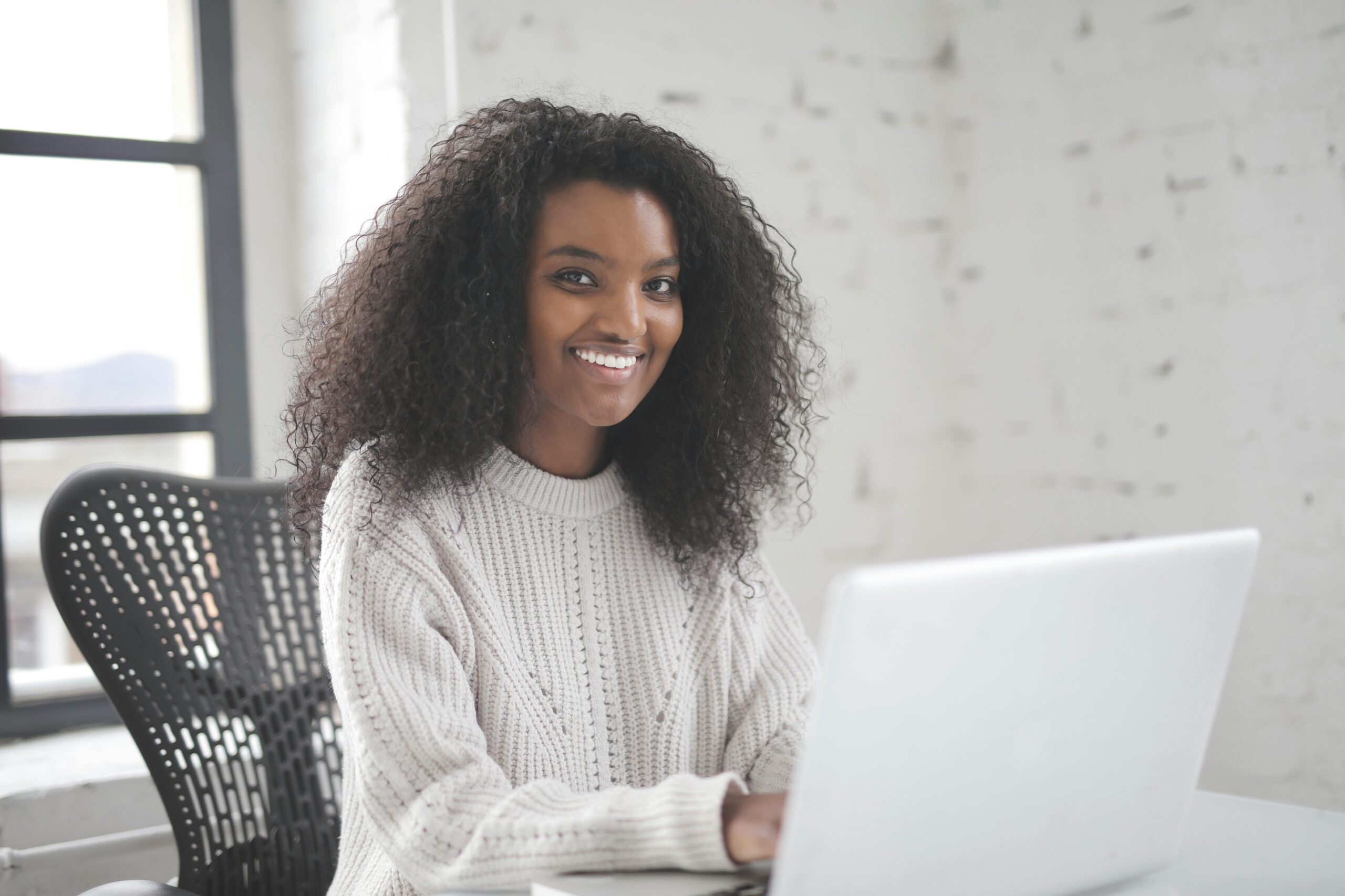 Smiling woman with curly hair using a laptop in a modern workspace, showcasing remote work confidence.